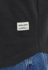 Crna majica od pamučnog materijala, s bijelom etiketom s natpisom "JACK & JONES", prišivenom na donjem rubu, u kombinaciji s plavim trapericama.