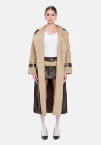 Trench-coat beige avec accents en cuir marron, ceinturé à la taille. En dessous, une mini-jupe en cuir marron. Le look est complété par un haut blanc et des bas.