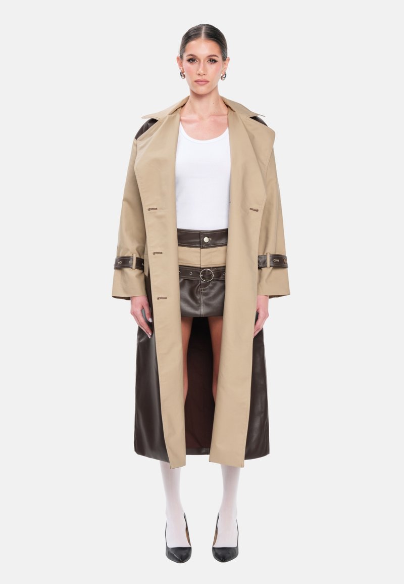 Trench-coat beige avec accents en cuir marron, ceinturé à la taille. En dessous, une mini-jupe en cuir marron. Le look est complété par un haut blanc et des bas.