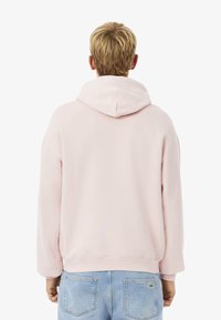 Sudadera rosa claro con capucha ajustable, puños acanalados y dobladillo. La parte posterior presenta un diseño simple sin marcas visibles. Combinada con jeans azul claro.