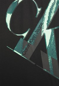 Black t-shirt featuring shiny teal and metallic silver graphics. The design includes the text "Calvin Klein Jeans" in a bold font.