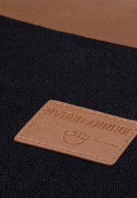 Barna bőr címke dombornyomott szöveggel fekete szöveten, varrott szegéllyel és vízszintes vonalakkal; minimalista dizájn, téglalap alakú.