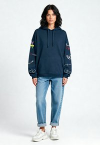 Marineblauwe, hoodie met geborduurde accenten op de mouwen. Gecombineerd met lichtblauwe, opgerolde spijkerbroek en witte sneakers.