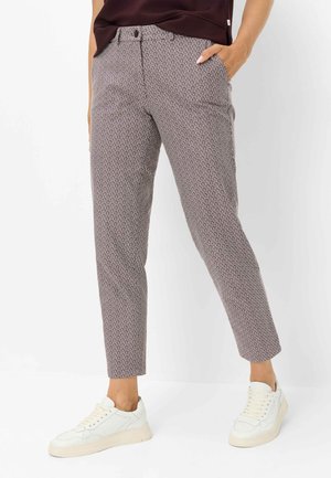 Personne portant un pantalon slim à motifs, main dans la poche et baskets blanches, debout devant un fond blanc uni.