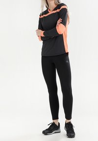 Maglia atletica nera a manica lunga con accenti arancioni, abbinata a leggings neri e calzature nere. Tessuto strutturato con una finitura testurizzata.