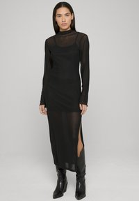 Robe noire à manches longues avec une superposition transparente, des fentes latérales et une silhouette ajustée. Portée avec des bottes noires montantes jusqu'au genou. Texture lisse.