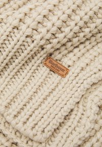 Pletená čepice z měkké krémové vlny s texturovaným vzorem. Obsahuje obdélníkovou koženou nášivku s embosovaným nápisem "Barbour England."