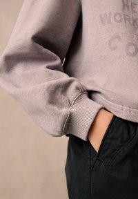 Felpa viola chiaro con polsini a coste e vestibilità rilassata, abbinata a pantaloni neri scuri. Mano visibile nella tasca, dettaglio del logo discreto.