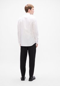 Balta long-sleeve krekli ar klasisku apkakli, taisna griezuma, ar vienu pogu aprocēm, valkāta ar melniem biksēm un melnām kurpēm.