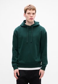 Ciemnozielona bluza z kapturem, wykonana z miękkiego materiału, z regulowanym kapturem, ściągaczami na rękawach i logo na klatce piersiowej. Nałożona na białą koszulę.