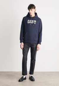 Felpa blu navy con logo bianco, tasca a canguro, cappuccio con coulisse, abbinata a jeans scuri e mocassini neri con calzini bianchi.