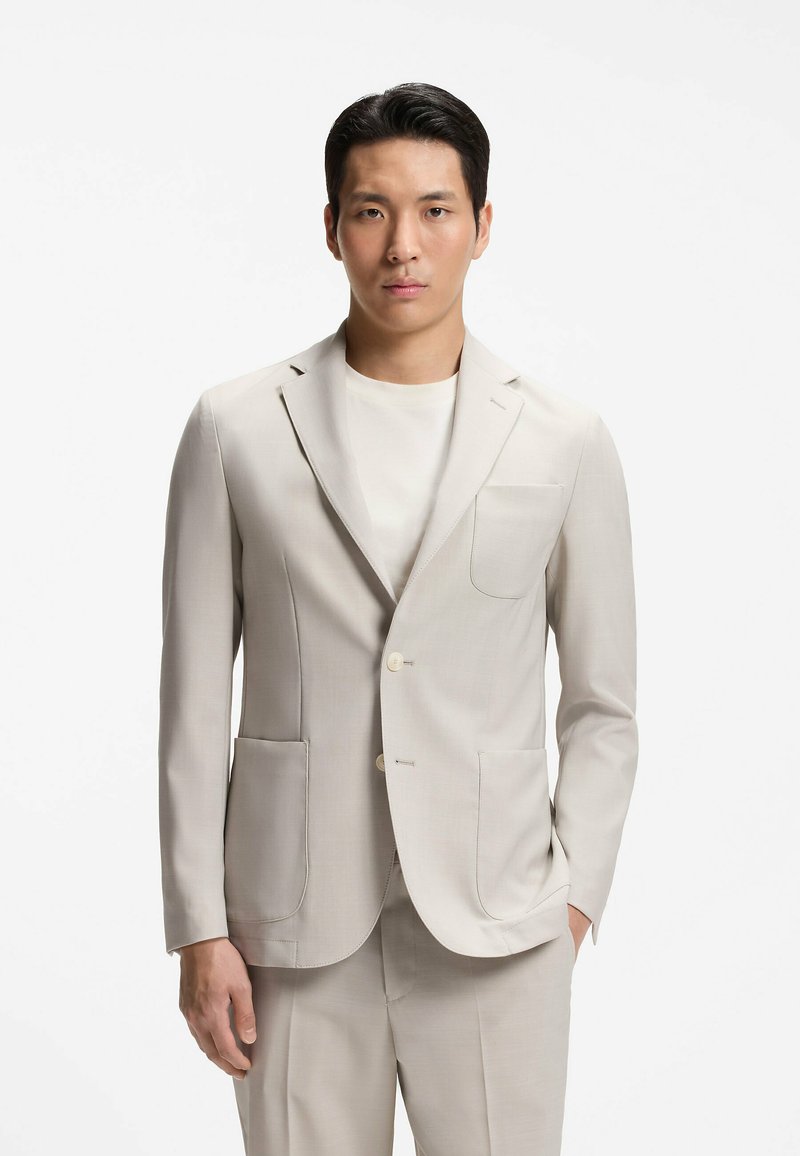 Homme portant une veste de costume beige clair et un pantalon assorti avec un t-shirt blanc à col rond, debout devant un fond blanc uni.