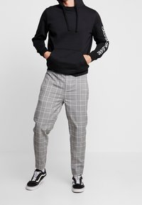 Černá mikina s kapucí, přední kapsou a stahovací šňůrkou, doplněná šedými kalhotami v kostkovaném vzoru s úzkým střihem. Outfit doplňují černo-bílé tenisky.