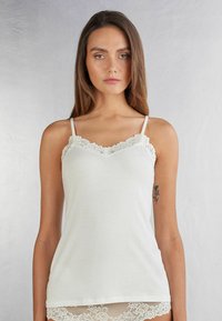 Vitt linne med spetskanter vid halsringningen, justerbara axelband och en mjuk, slät tygstruktur. Klassisk och enkel design.