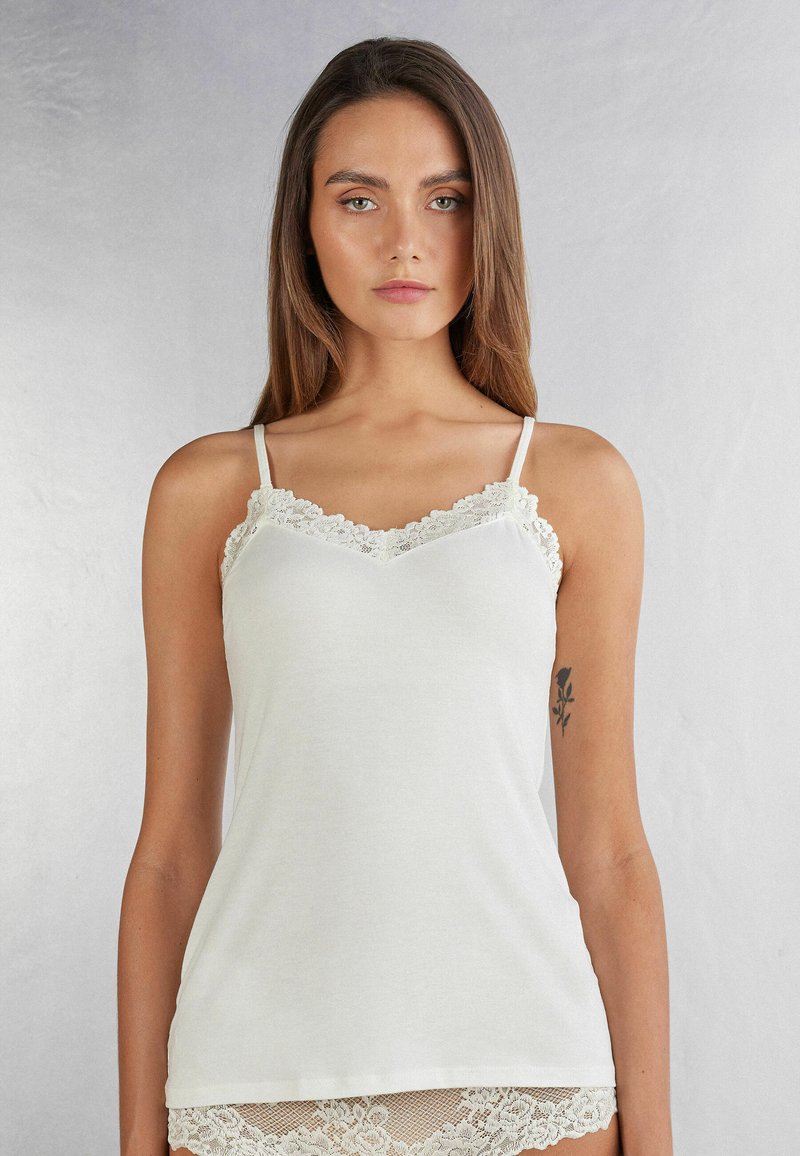 Vitt linne med spetskanter vid halsringningen, justerbara axelband och en mjuk, slät tygstruktur. Klassisk och enkel design.