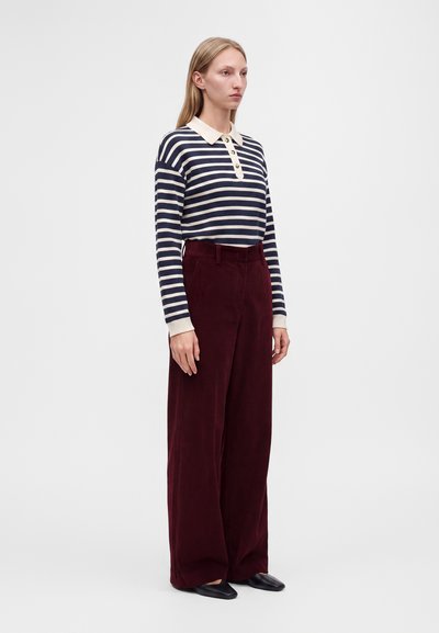Femme debout portant un polo à manches longues rayé bleu marine et blanc et un pantalon large taille haute rouge foncé avec des chaussures noires.
