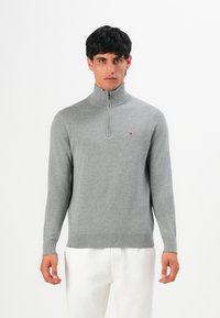 Mann in hellgrauem Quarter-Zip-Pullover und weißen Hosen, der vor einem schlichten weißen Hintergrund steht und nach vorne blickt.