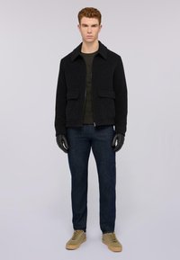 Svart ullblandad jacka med krage, två framfickor, bärs över en mörkgrön tröja, kombinerad med blå denimjeans och handskar.