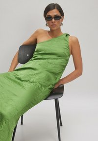 Grünes One-Shoulder-Kleid mit gekräuselter Textur, das eine schlanke Silhouette hat. Modell sitzt auf einem schwarzen Stuhl und trägt dunkle Sonnenbrillen.
