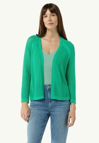 Grüne gestrickte Strickjacke mit offenem Vorderteil, langen Ärmeln und strukturiertem Muster, über einem gestreiften Tanktop getragen und kombiniert mit blauen Jeans.