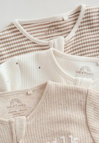 Trzy miękkie, neutralne tonalnie bluzeczki dla niemowląt z delikatnymi paskami i małymi kropkami, starannie ułożone warstwowo z widocznymi metkami marki NEXTbaby.