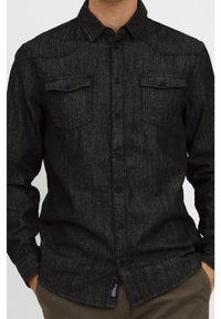 Musta deniimapaita, jossa on teksturoitu pinta, sisältää teräväkärkisen kauluksen, kaksi rintataskua ja edessä napitus.