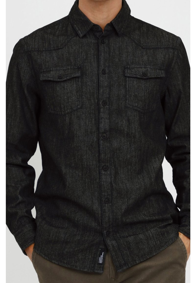 Musta deniimapaita, jossa on teksturoitu pinta, sisältää teräväkärkisen kauluksen, kaksi rintataskua ja edessä napitus.