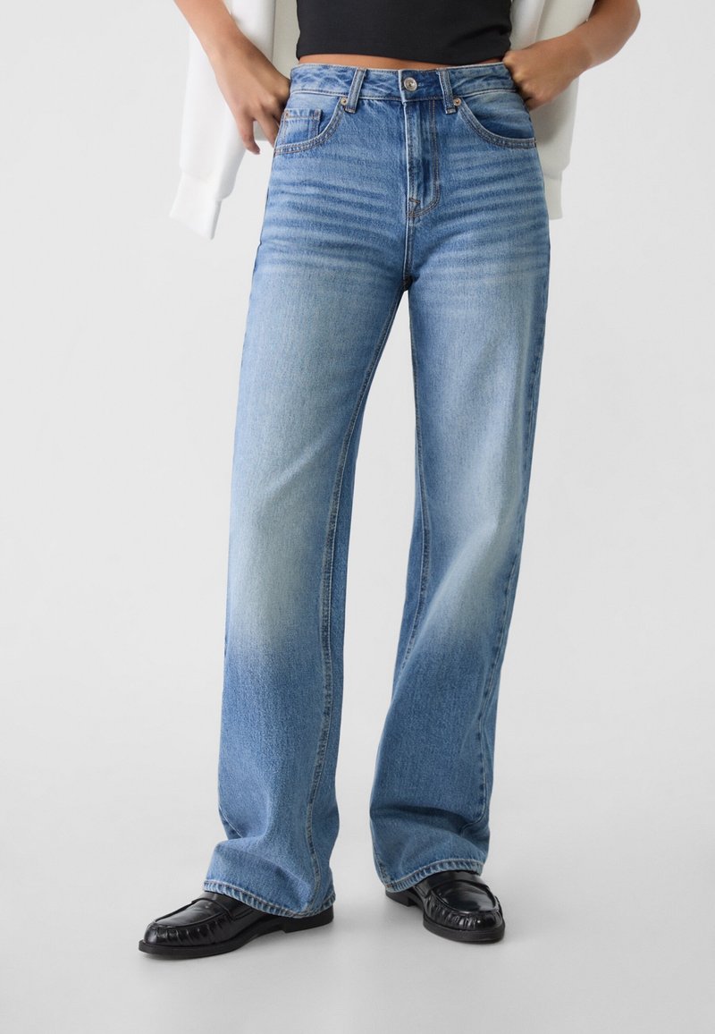 Jeans de denim de color azul claro, de pierna ancha, con zonas desgastadas, cinco bolsillos, cierre de cremallera y botón. Combinados con calzado negro.