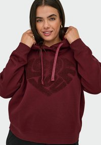 Burgundy huppari, jossa on teksturoitu sydänkuvio. Varustettu kiristettävällä hupulla, pitkillä hihoilla ja rennolla istuvuudella. Pehmeä kangas.