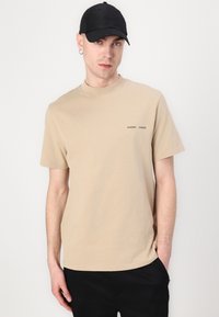 Beige színű pamut póló kerek nyakkivágással és rövid ujjakkal, amelyen egy kis fekete márkájú logó található a mellkason. Fekete nadrággal és fekete sapkával viselve.