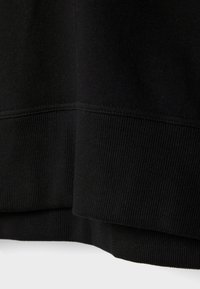 Détail du tissu d'un sweat-shirt noir montrant les poignets et l'ourlet côtelés en gros plan sur fond uni.