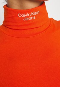 Orange turtleneck-top av mjukt material, med vit text "Calvin Klein Jeans" på kragen. Slät yta med en figursydd design.