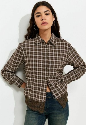 Young woman with long dark hair wears brown plaid button-up shirt with lace trim and blue jeans, standing with hands on hips.