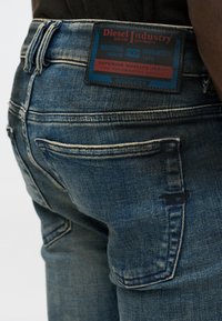 Jeans de denim azuis com um rótulo de marca visível na cintura traseira, design de cinco bolsos e detalhes de costura em contraste.