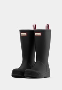 Fekete gumicsizma sima felülettel, piros és fehér csíkos húzófülekkel, valamint téglalap alakú márkajelzéssel az oldalon.