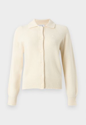 Femme aux longs cheveux noirs portant un gilet boutonné jaune pâle, un jean bleu clair à jambes larges et des baskets blanches sur fond blanc.