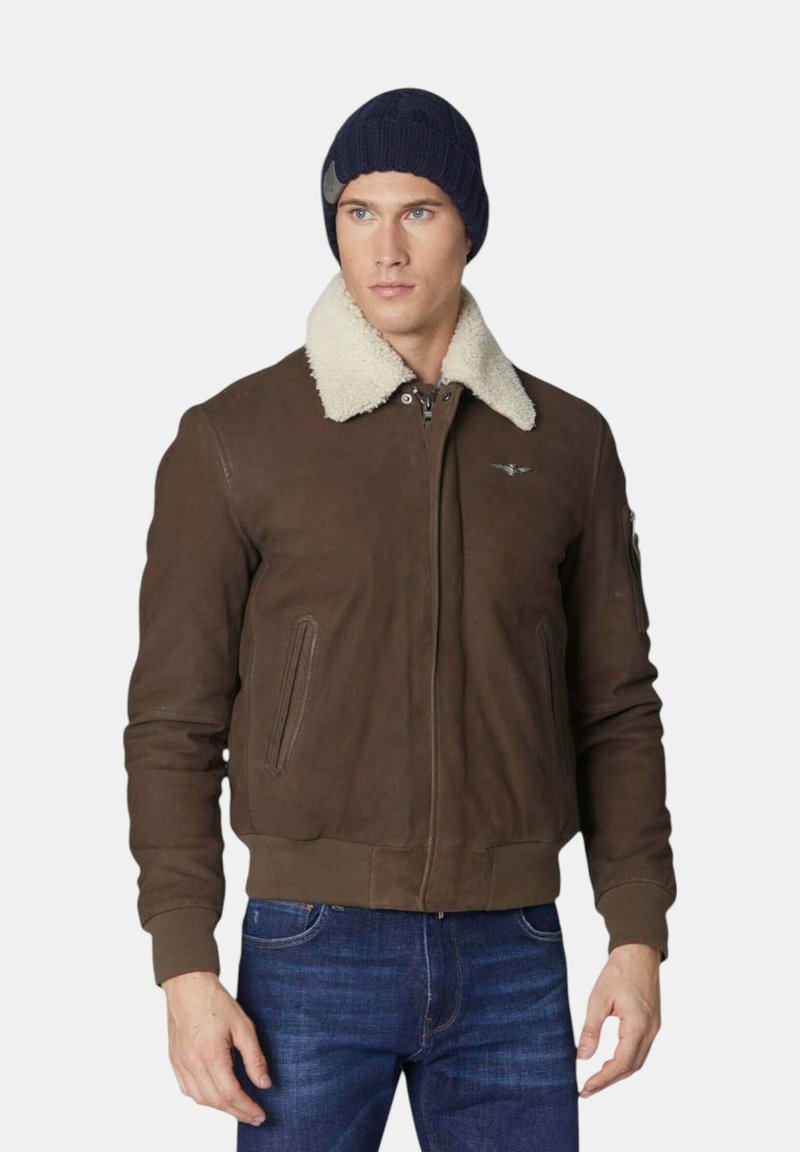 Bruine suedela jacket met een schapenvachtkraag, ritssluiting aan de voorkant, zijzakken en een logo op de borst, gecombineerd met een marineblauwe gebreide beanie en blauwe spijkerbroek.