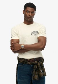 Beige crew-neck t-shirt with black graphic "Tour 1979" print; worn with a plaid flannel shirt tied at the waist and blue jeans. Gold watch on wrist.