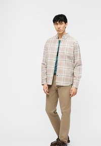 Νεαρός άνδρας που στέκεται, φοράει μια μπεζ πουκαμίσα με καρό κουμπιά πάνω από μια τιρκουάζ μπλούζα, χακί παντελόνι και σκούρα καφέ παπούτσια σε λευκό φόντο.