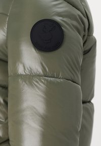 Giacca a vento imbottita verde oliva con una texture liscia e lucida, con una toppa in gomma sulla manica che raffigura un'anatra e la scritta "SAVE THE DUCK."