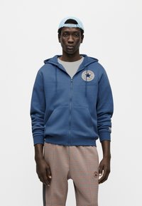 Μπλε φούτερ με φερμουάρ και τσέπη καγκουρό, με κυκλικό λογότυπο Converse στο στήθος. Συνδυασμένο με καρό παντελόνια.