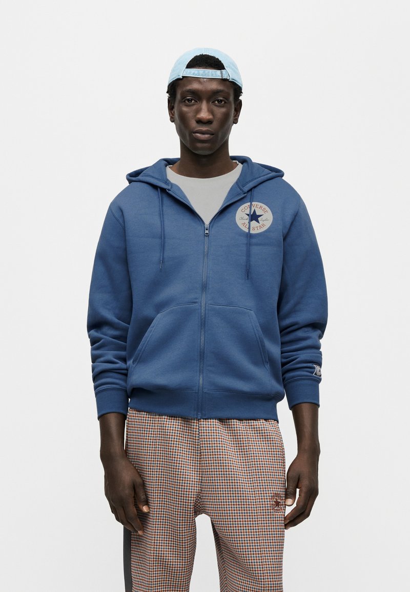Blå hættetrøje med lynlås og kængurulomme, med en cirkulær Converse-logo på brystet. Parret med ternede bukser.