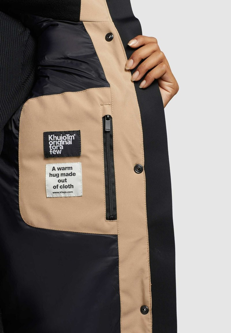 Main dans une veste beige et noire ouverte, montrant une poche intérieure avec deux étiquettes en tissu et une fermeture éclair.