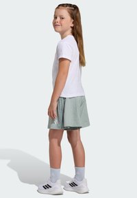 White short-sleeve t-shirt, light green skort with polka dots, white sneakers with black stripes, light blue socks. Child facing slightly sideways.