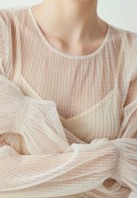 Przezroczysta, kremowa bluzka z pionowym, teksturowanym wzorem, miękkim, okrągłym dekoltem i długimi, luźnymi rękawami. Widoczna warstwa pod spodem. Delikatna, lekka tkanina.