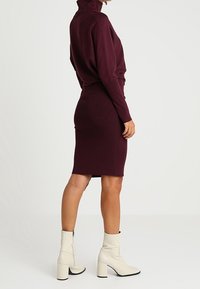 Donna che indossa un vestito a maglia bordeaux attillato e stivaletti bianchi alla caviglia, in piedi di lato contro uno sfondo chiaro e semplice.