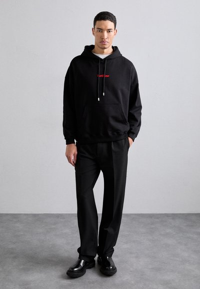 Črna kapuca sweatshirt s rdečim logotipom, kenguru žep, rebrasta manšeta; kombinirana s črnimi krojenimi hlačami in sijajnimi črnimi čevlji.