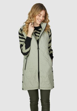 Gequilted vest in lichtgroen met een hoge kraag, zwarte ritsen en drukknoopjes aan de zijkanten. Gecombineerd met een gestreepte zwarte en groene trui.