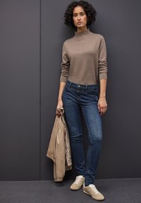 Beige stickad polotröja, mörkblå smala jeans, håller en ljusbrun jacka. Ljusfärgade sneakers med vita snörningar. Lockigt hår.