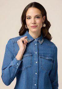 Cămașă din denim cu guler împodobit cu perle, cu nasturi pe față și două buzunare pe piept, într-o nuanță medie de albastru. Mâneci lungi cu manșete cu nasturi.